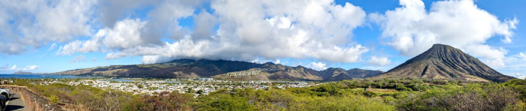 a panoramic view at hawaii kai lookout on the windward coast. Spanning from far off in the distance on the left is diamond head crater, which appears very small now. There are towns in the middle, with the taller mountain range behind. These are much taller because they are already in the clouds in the sky above. Continuing to the right you end with koko crater mountain. 