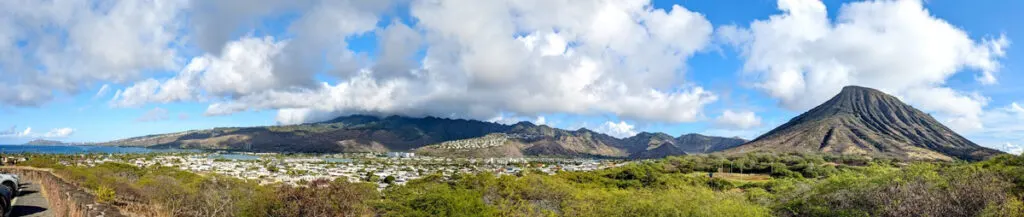 a panoramic view at hawaii kai lookout on the windward coast. Spanning from far off in the distance on the left is diamond head crater, which appears very small now. There are towns in the middle, with the taller mountain range behind. These are much taller because they are already in the clouds in the sky above. Continuing to the right you end with koko crater mountain. 