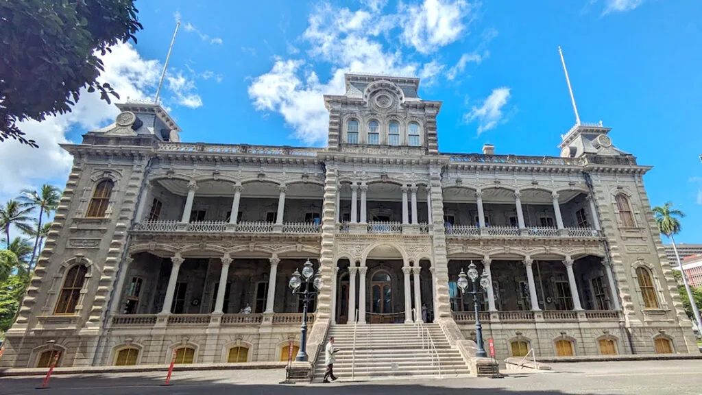 an old style building from the 1800s, but its confused on the style, it looks baroque but isn't beautiful. It's very grey and cement like. It has a tower in the center, with covered balconies on either side. The sky above it is bright blue with a few scattered white clouds. This is Iolani Palace in the middle of downtown honolulu