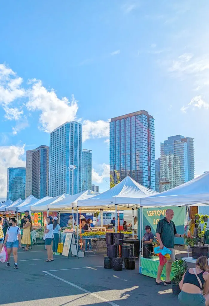 kaka'ao farmers market in honolulu. The image showcases the contrast of new and old. Pop up tents with white covers and poles holding them up for shade, stand in a nice row lining the image from front to back. People are gathered at each stall looking at the produce. In the background, towering above are high rise buildings. 