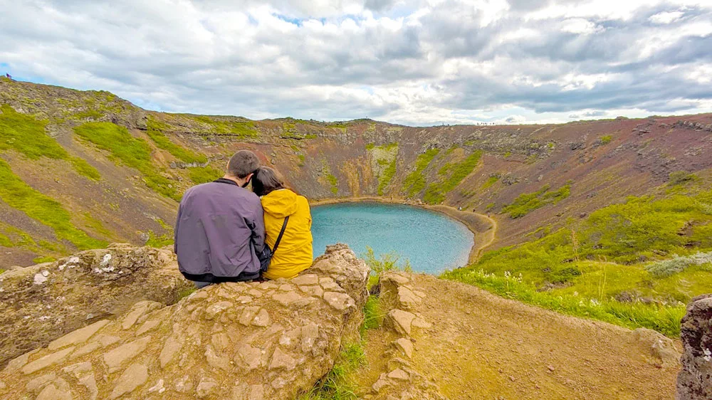 a couple sits on a brownish red rock ledge. Around them it's dirt and some green grassy patches. The dirt and grass shapes a circle going downward into a crater, with blue water in the centre. The sky is ominous, with dark clouds but you can still see some blue spots between the clouds.