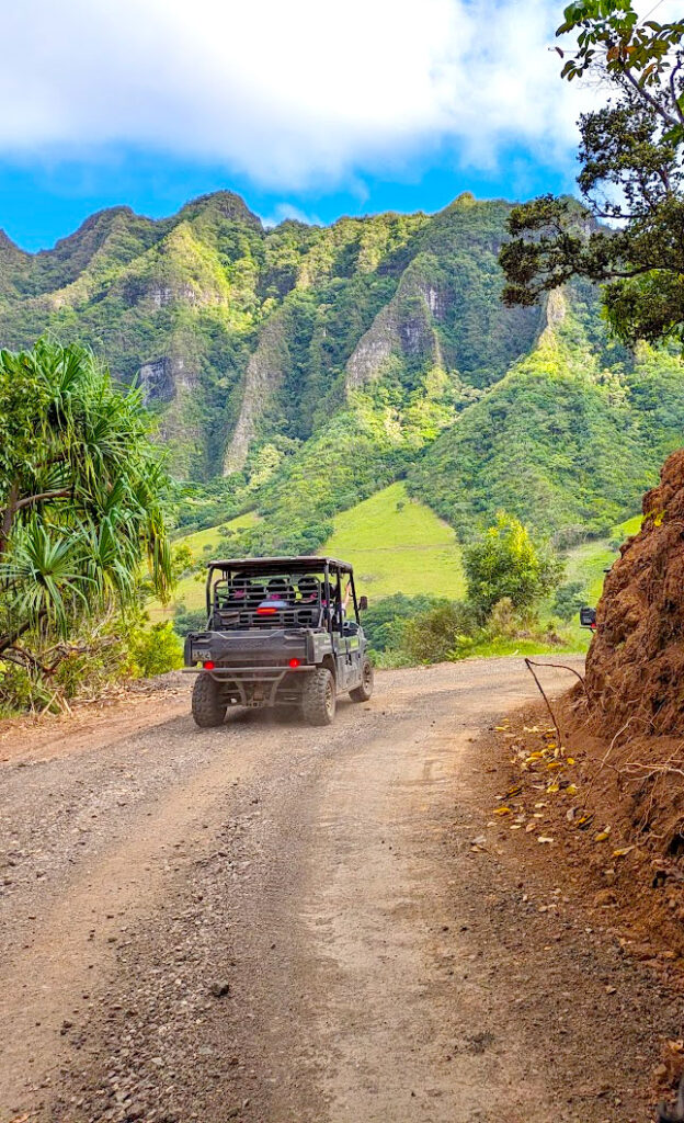 driving through jurassic world. On a dirt road, there's a UTV ahead creating dust behind it. They're turning a corner. Off the road are the soaring kualoa mountain range. There's a line of blue skies before white clouds take over on the top of the image. 