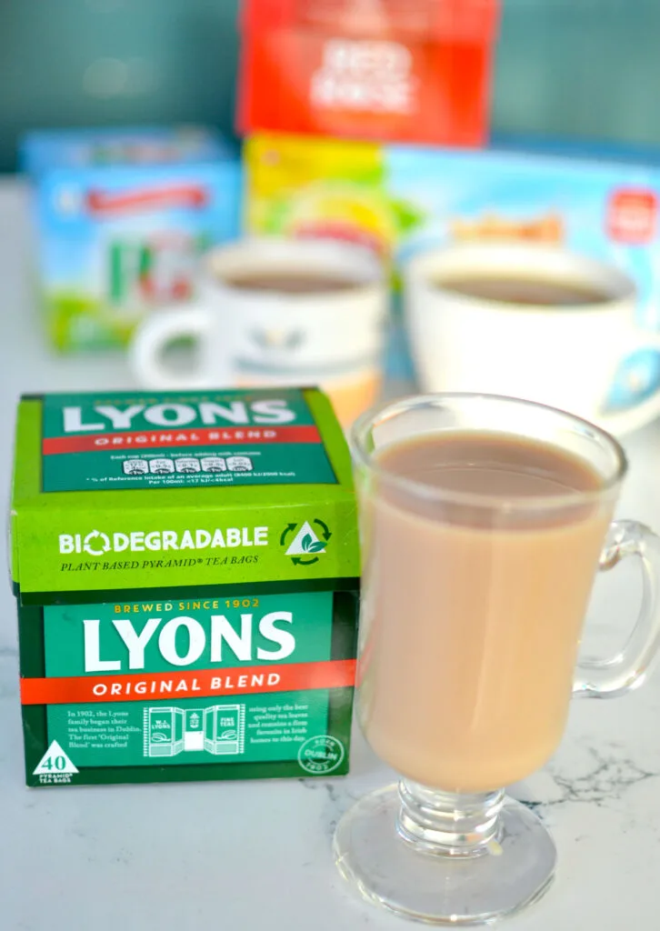 a vertical image with an irish style glass mug that shows a brewed tea with milk inside. Next to the mug is the bag of tea for LYONS original blend tea. Blurred in the background are two other mugs with tea, and three other tea boxes. 