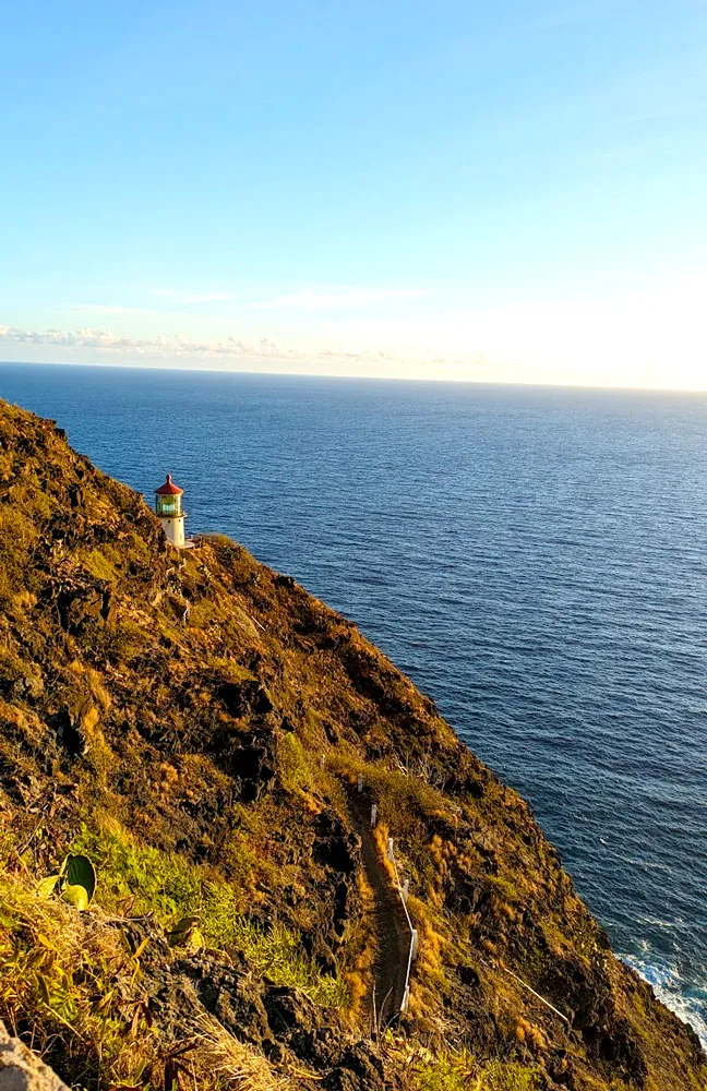 a stark but simple image of a golden cliffside with the sun shining on it during sunrise. There's a small white lighthouse with a red roof that steels your focus. The mountain is against the vast ocean that leads into the sky.