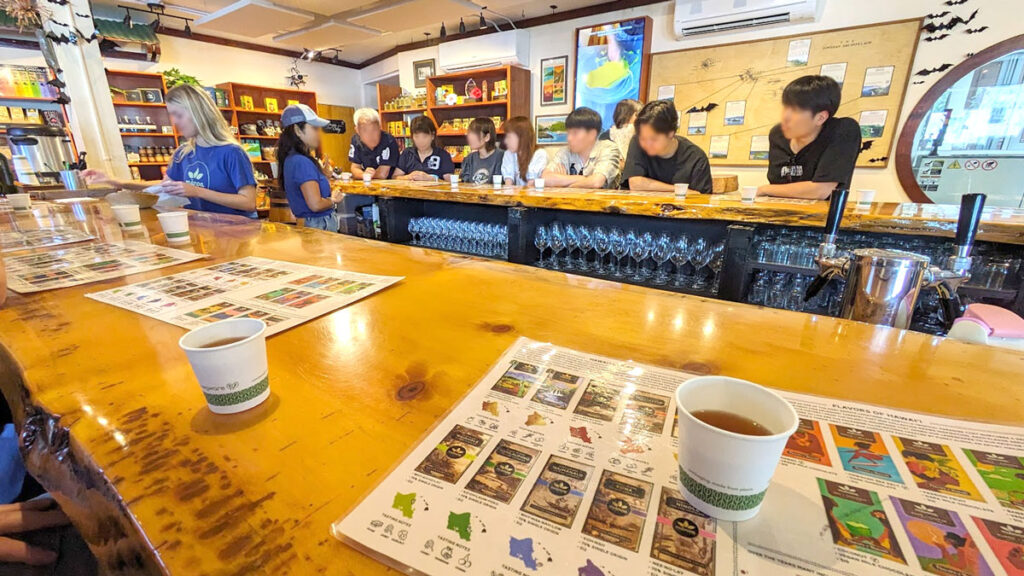 manoa chocolate tasting. Sitting at a long bar, there are sheets in front of people at the bar. On the sheet is a list of manoa chocolate bars and their tasting notes. You can see about 3 on the table, shared between two people each. There are also small paper cups filled with a brown liquid. Two people stand behind the bar, one is facing your table. The other is facing the opposite bar on the other side, where you see a group of six people doing another tasting. This is in the middle of the manoa chocolate store.