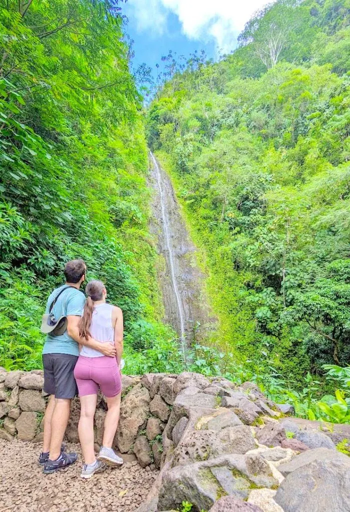 a couple stand in workout attire, with their backs to the camera. The mans arm is wrapped around the woman's back. They are both looking at a small waterfall stream that's over rocks, but surrounded by green lush vegetation. You can see a snippet of the sky above. 