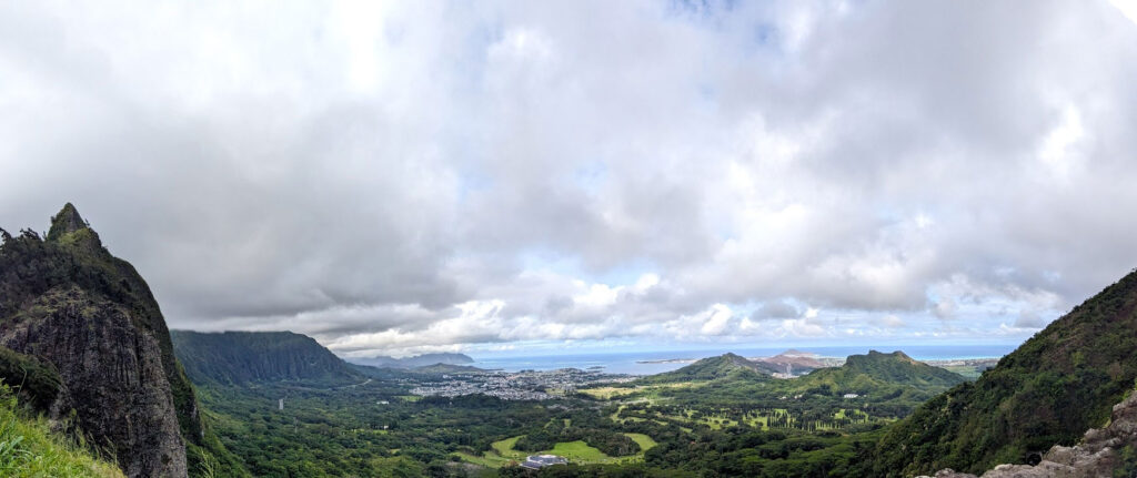 nuuanu lookout point on the windward coast or actually of the windward coast. The right and left sides of the photo are bordered by mountains ranges. they lead you down into the town below abd finally the ocean beyond it. There are thick white and grey clouds in the sky above. 