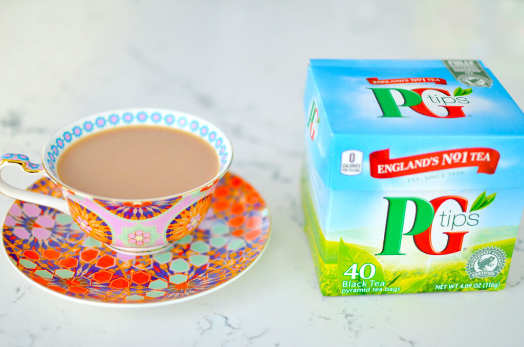 a delicate teacup with a purple red and green geometric floral pattern on it. It's filled to the brim with tea and a dash of milk added. Next to the teacup is the closed box for PG tips tea bags.