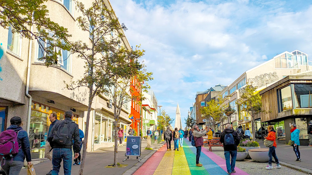 a rainbow stripes on the pavement street lead your eye to the end where a steeple of a church stick out above the pedestrians walking in front of it. On either side of the street are buildings with small trees.