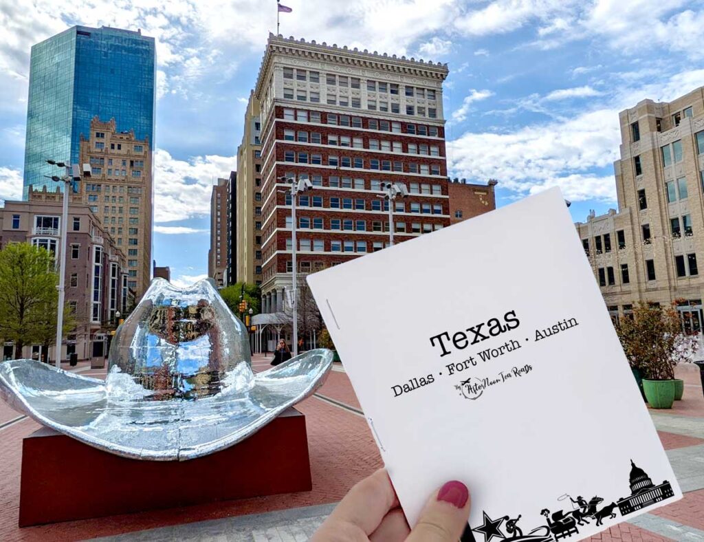 a hand holes up a printed pocket booklet that isn't much larger than her hand. It says Texas in large, with Dallas - Fort Worth - Austin in a smaller font below. There's a black and white Texas graphic lining the bottom.  In the background is a large disco cowboy hat with a city view of a downtown area.