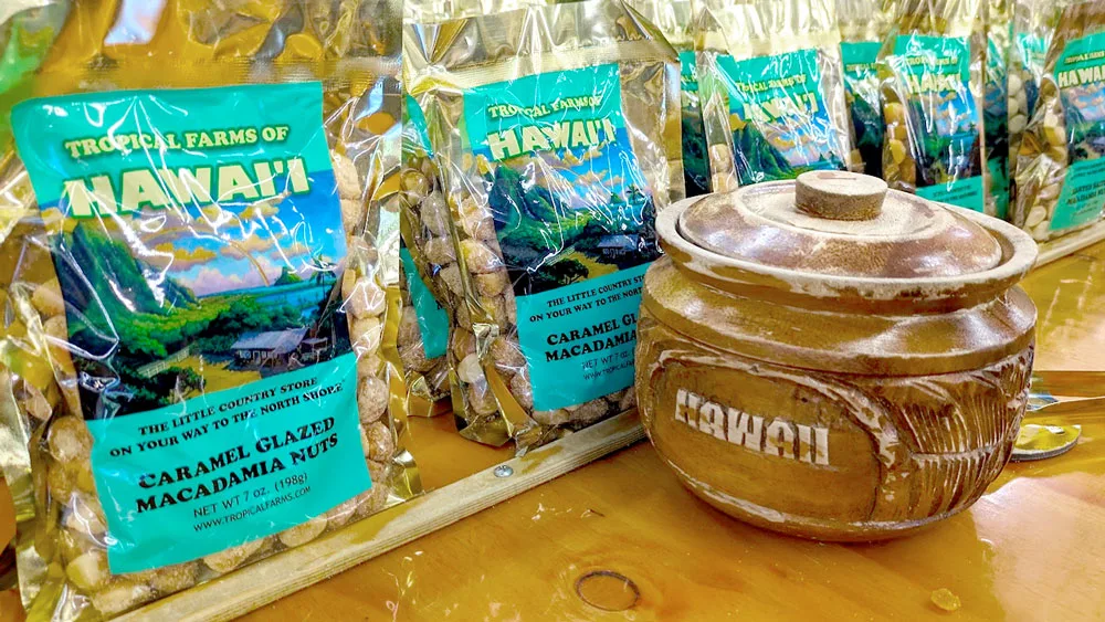 a closeup of several rows of bags of macadamia nuts from Tropical Farms of Hawai'i. The label is a bright teal colour and a landscape painting of hawaii in the middle. You can see the macadamia nuts through the clear bag. The closest flavour is caramel glazed macadamia nuts. There's also a bowl on the table with a lid. The bowl is filled some free tasters.