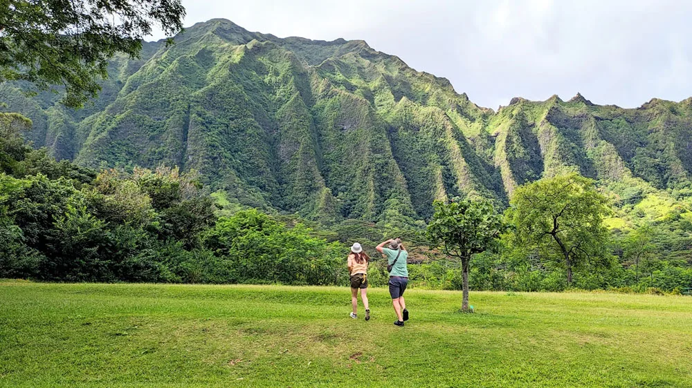 two people running away with their back to the camera in the middle of a field while giant mountain ranges loom above them in the clouds