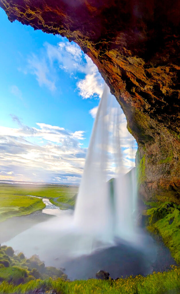 Seljalandsfoss waterfall day trip from reykjavik at golden hour. You're standing behind a waterfall that pouring over from a cliff above, it's all lit up yellow form the setting sun. Creating a beautiful calming golden hue to the phot on the rocks, and the mist in the air from the waterfall. You can see a valley in the distance, and the sky is a blue and yellow as the sun starts to set.