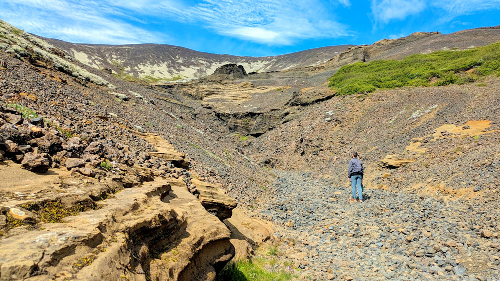 a man stands in the middle of a rocky path... that isn't really a path at all. It leads further into the rocky mountains of Iceland. It's all different shades of brown, with a little bit of mossy grass that has grown in the corner. The mountains meets the blue sky with whispy white clouds. No one else is around. 