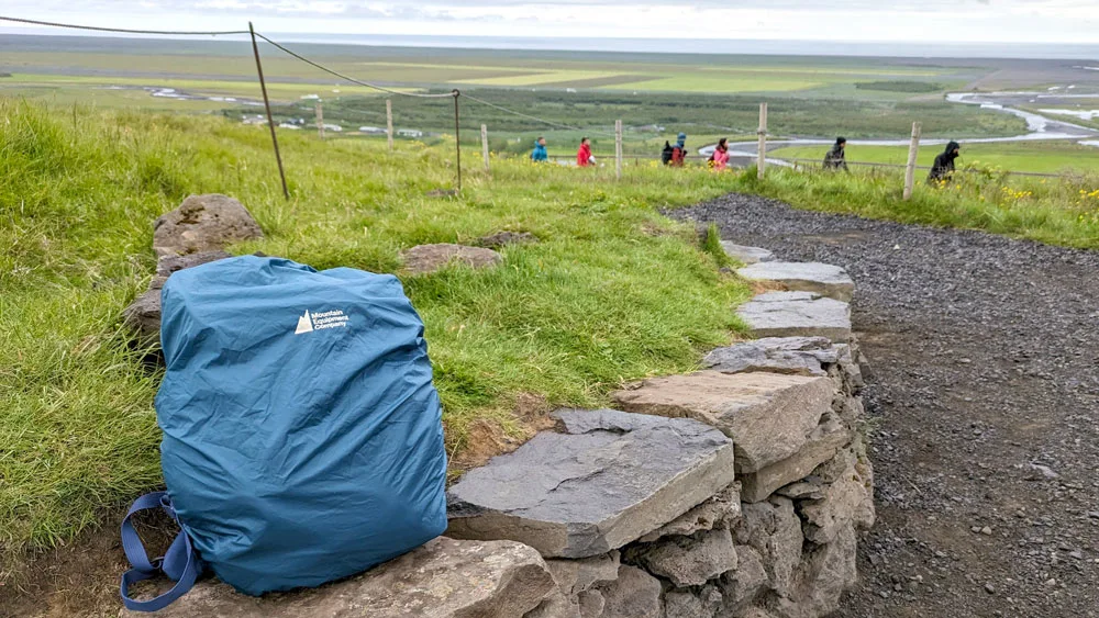 a lbag that's covered in a rain cover sits on a stone wall against a grassy hill. It's on the side of a path, you can see people walking up in the distance, and below them is the Icelandic valley with a stream going the whoel way to the ocean. You can see the ocean even meet the horizon. It's a grey day. 