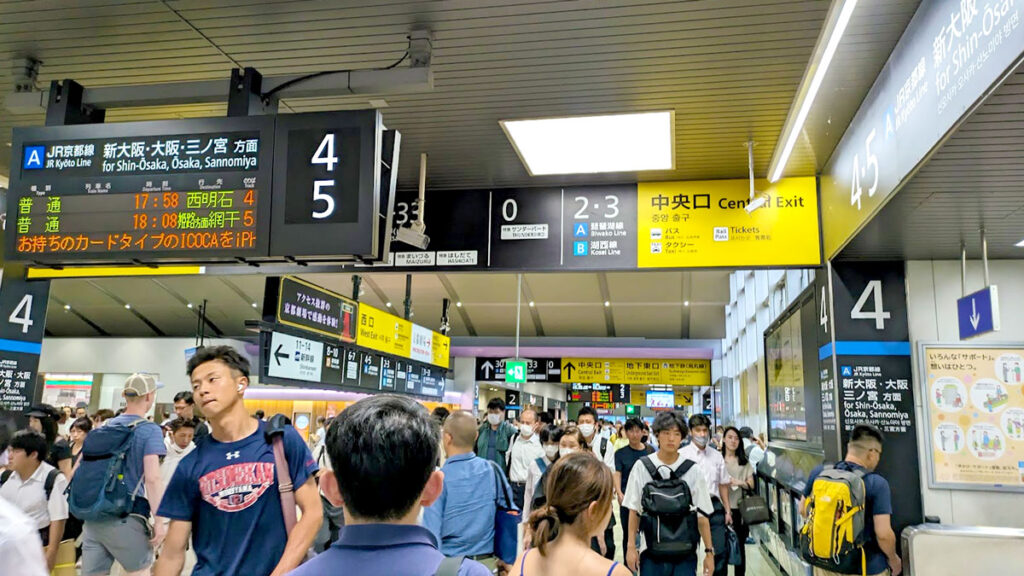 a sea of people walk in every direction in a train station. Above the people is a mess of signs, there ar a lot of them telling you where you need to go. The signs are a mix of japanese and english 