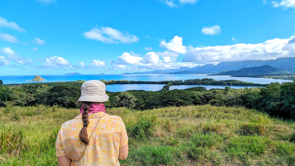 a woman stands with her back to the camera. She looks like an explorer, wearing a yellow floral collared shirt. She has a pink neck gaiter and a beige bowl hat. You can see her brown hair on her back, in a braid. In front of her is the vast Hawaiian view of the ocean, with a distinct island not too far from shore - mokolili island (or china man's hat, or the little lizard). In the distance, you still the start of the mountain ranges, growing larger as they go more in land. They are cascading, so they also get lighter the farther they go from the viewer. clouds are leading off the mountains, separating over the ocean, leaving more blue skies visible.  