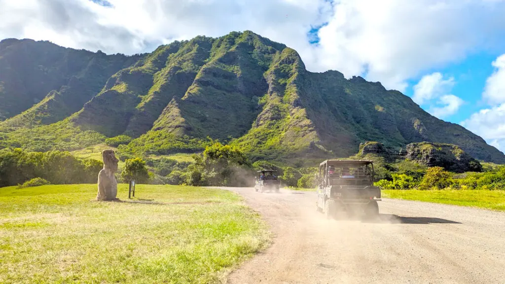 two utv driving in a line on a dirt road leaving a dusty mist in their wake. There's lush green grass on either side of the road, with an old tiki looking stone statue looking at the road. The cars are driving towards a large cascading grassy mountain range. The sky is a mix of blue skies and white and grey clouds above the mountains.