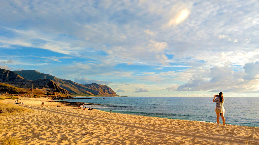 the gorgeous golden hour on the leeward coast at yokohama bay on oahu hawaii. the perfect end to a one week in O'ahu. A woman stands on the right taking a photo with her phone of the vast rocky mountains that wrap around the beach beach and the ocean meeting the shore. 