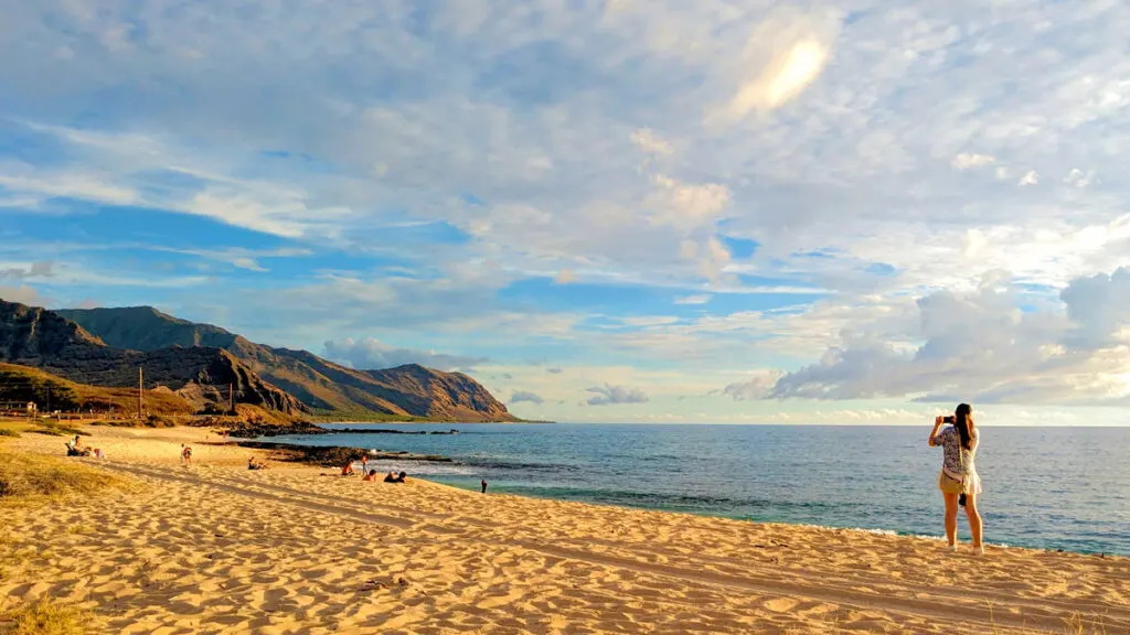 the gorgeous golden hour on the leeward coast at yokohama bay on oahu hawaii. the perfect end to a one week in O'ahu. A woman stands on the right taking a photo with her phone of the vast rocky mountains that wrap around the beach beach and the ocean meeting the shore. 