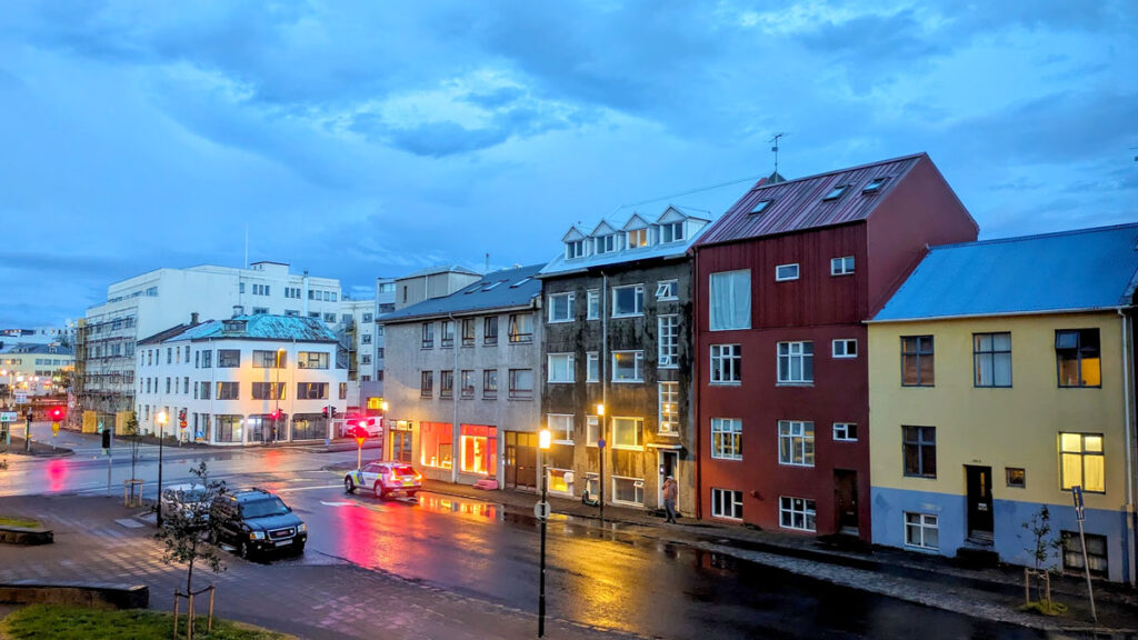 a view outside a hotel room at 12:38 a.m. in Iceland in the summer. It's dusk, with a blueish hue across the photo. One car is driving, stopped at the red light on the street. Otherwise the city is quiet. Street lamps are on, offering a yellow touch the image. And there's a reflection of the lights on the wet pavement below. The street is four buildings baout three to four stories each that are all attached, but also all different colours. From beige, to brown, to red, and yellow. 