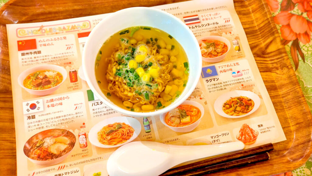  a top down view of a japanese ramen menu with pictures of different ramens you can order. There's a mini ramen bowl on top of it, with noddles corn and green herbs. There's an upside down ramen spoon below it. There's an orange hue to the image from the lighting in the restaurant.