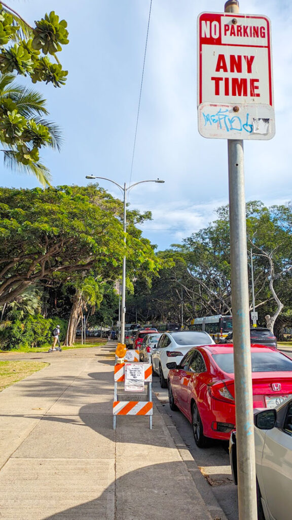 a vertical photo showing the sidewalk with cards parks all along side it. There's a pole on the right, at the top is a sign that is red and white. It reads: No Parking ANY TIME. There's another section below that looks like it was painted over and has blue graffiti on top, but you can slightly see the ENDS underneath it. 