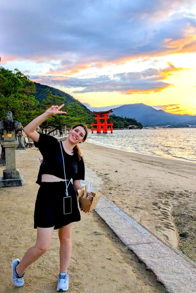 learning a universal sign in japan. A woman poses in a picture with a big smile on her face and her arm is up in the arm waving a peace sign with her hand. She's walking on the shore , there are stone lanterns lining her walk behind her, leading to a large red tori gate in the middle of the water. Large mountains cascade through the background as the sun starts to set behind them