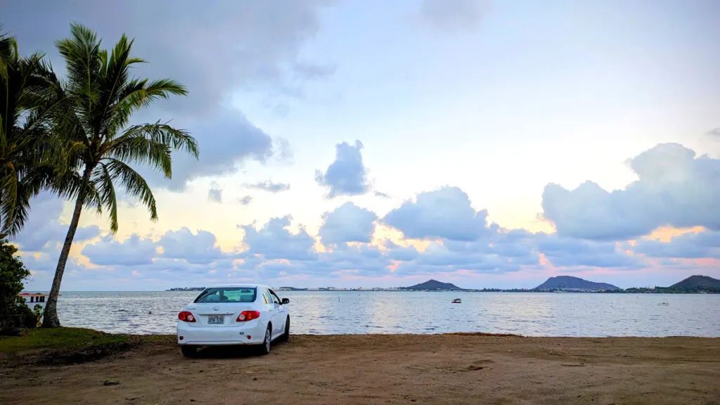 the perfect sunset in O'ahu. A white toyota corolla is parked on the edge of the sand. it looks like the water is right underneath it, that's how close it is to the edge. There's a hanging palm tree on the left of it. But otherwise it's an empty sand dune. The water leads off into the sunset sky of pastel pink, yellow and blues with some clouds over the horizon and some mountains. 