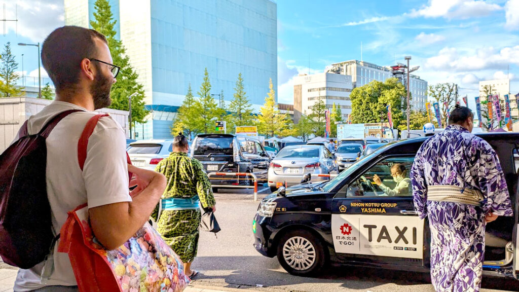 a man standing on the left side of the phot, with a backpack and large reusable shopping back. He's looking at a much larger man in a kimono and his hair in a top bun, he looks like hes getting into a taxi. You know it's a taxi because in English, it's written TAXI  in English 