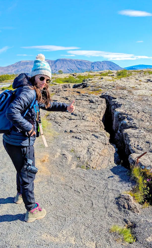 a woman stands in front of a crack in the ground (that goes all the way down) in Iceland in the summer. It's a sunny day with basically clear skies. Theres a mountain (that's probably a volcano) on the horizon. The woman is wearing black jogger pants with hiking boots, a puffer jacket, and a hat and sunglasses.