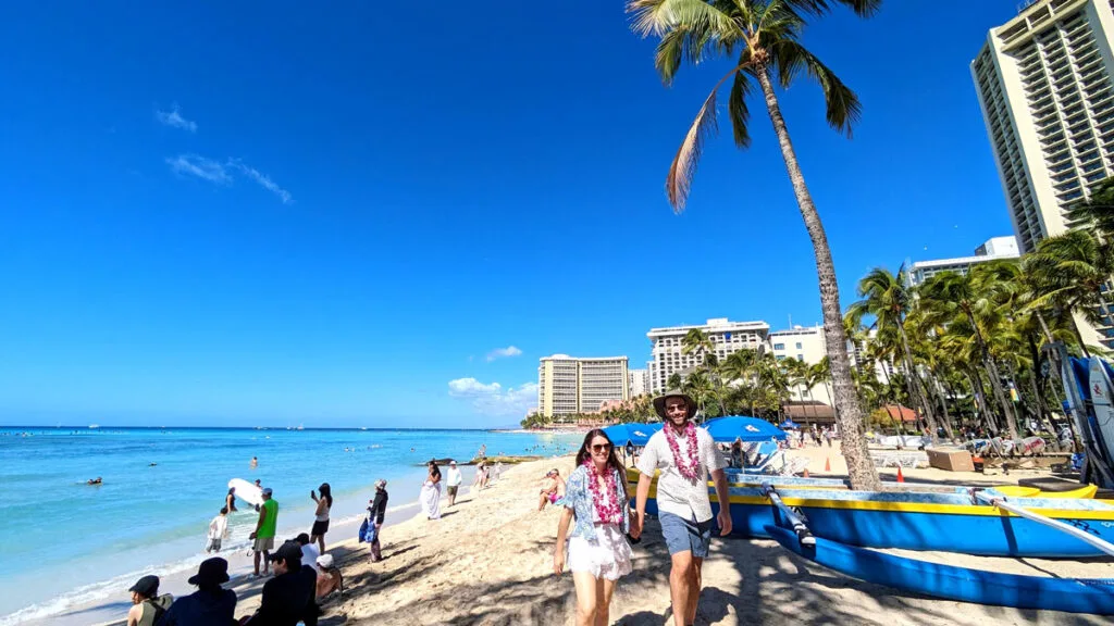 a couple is at the foreground of the photo walking towards the camera about to spend one week in O'ahu. They're holding hands, both wearing matching purple leis, and outfits... the woman wears a white skirt with a white tank top and a blue open blouse on top. The man wears a white button down shirt and blue shorts. The beach is busy, there's an outrigger next to them and one sole palm tree. But there are more palm trees lining the end of the beach, before the tall hotel buildings take over the skyline. The water in the ocean is a beautiful clear blue, matching the sky above. 