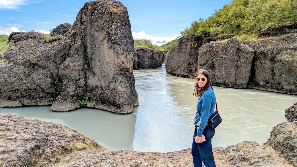 a woman stands in front of a pool of water that's carving the rocks as it goes by/ She's wearing sunglasses, pants, and a thin quarter zip sweater - no jacket in the afternoon in the summer in Iceland is needed!