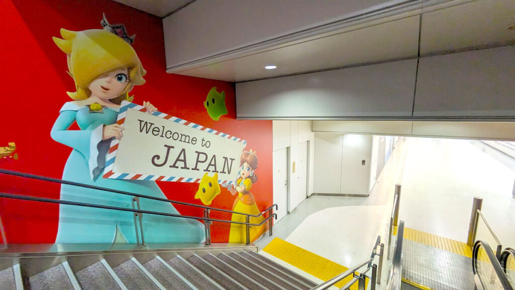 going down the escalator, there are stairs  next to a wall with a mural painted on it. The background is red. There's a giant Daisy on the left, and smaller princess peach at the bottom of the stairs. They are both holding up a sign: Welcome to Japan - that's only in English and no Japanese