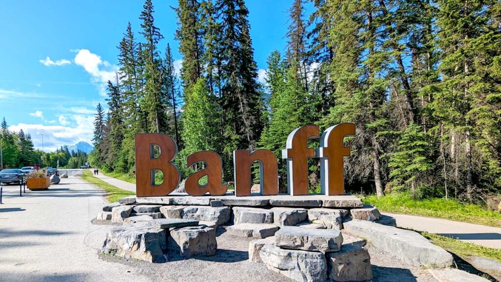 brown letters standing up to read: Banff  on the side of the road as you entre the town. There's a mountain in the distance, and trees to your right.