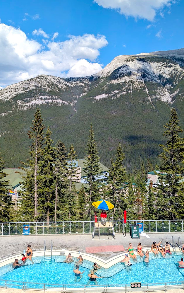 a view overlooking the banff upper hot springs that are overlooking mount rundle in the canadian rockies. It's a bum shaped pool (two half circles) with people sitting along the edge. It looks like a typical swimming pool There's one lifeguard in a high seat, with a blue and yellow iconic old school umbrella shading the lifeguard. The mountains are the view everyone is looking at, rocky mountains covered in green trees before the sky takes over the top third of the photo with a giant cloud passing above on an otherwise blue sky and sunny day.