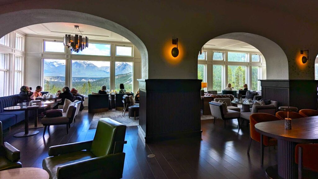 a dark room with cushy armchairs and tables. There. are two archways into another section of the room, where people are seated near the large windows behind them. This is why its so dark inside. The windows are the perfect viewpoint of the bow river, and the canadian rockies.