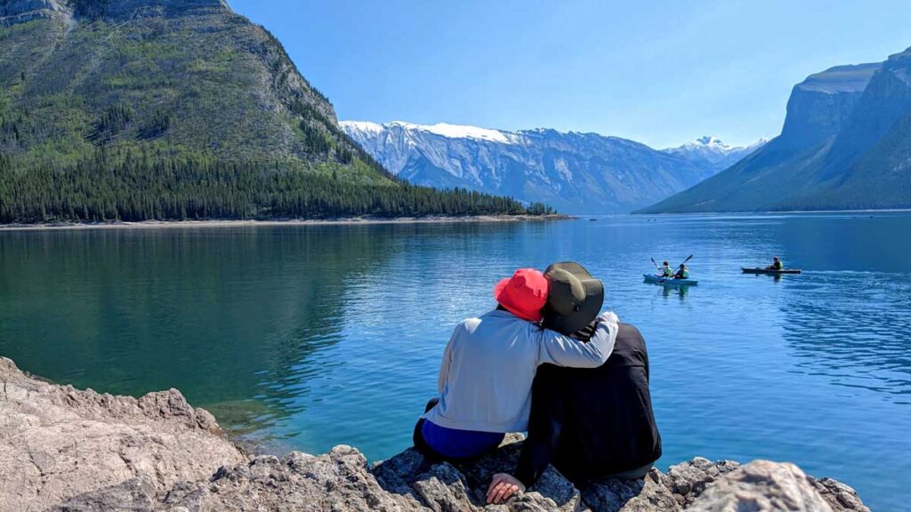 the back of a couple that sits on a rock overlooking lake minnewanka. There are people kayaking on the water. The lake leads all the way into a valley of mountains in the canadian rockies.