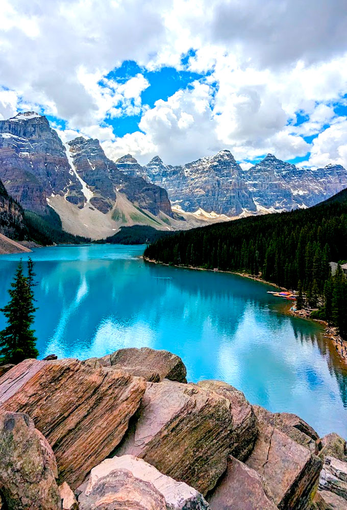 the vibrant blue water at moraine lake form the rock pile. Rocks lead your eye to the lake from the bottom of the photo, jutting out from the ground. The lake is so bright but still able to reflect the trees around it, the sky, and the mountains in the distance at the far end of the lake. They are in a row leading across the entire photo.