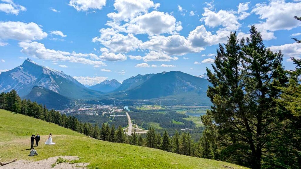 the view of mount norquay lookout. It looks like a large grassy field, but that's a mountain's edge. It slowly dips down, towards the view of banff town below and the rocky mountains in the distance. 