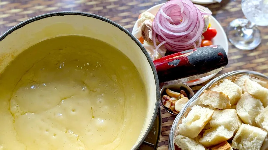 an overhead photo of a pot of cheese fondue with pieces of bread in a bowl and vegetables next to it, although you can really only see a pile of onions on top.