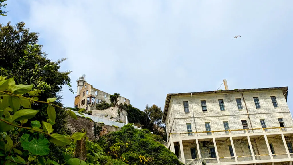 The view looking up the rock wall on alcatraz. There's a white painted brick building on the right, further up the hill is a ruined building, and you can see a small bit of the concrete lighthouse. The sky is overcast, but you can slightly see blue skies behind the cloud and there's a seagull flying across.