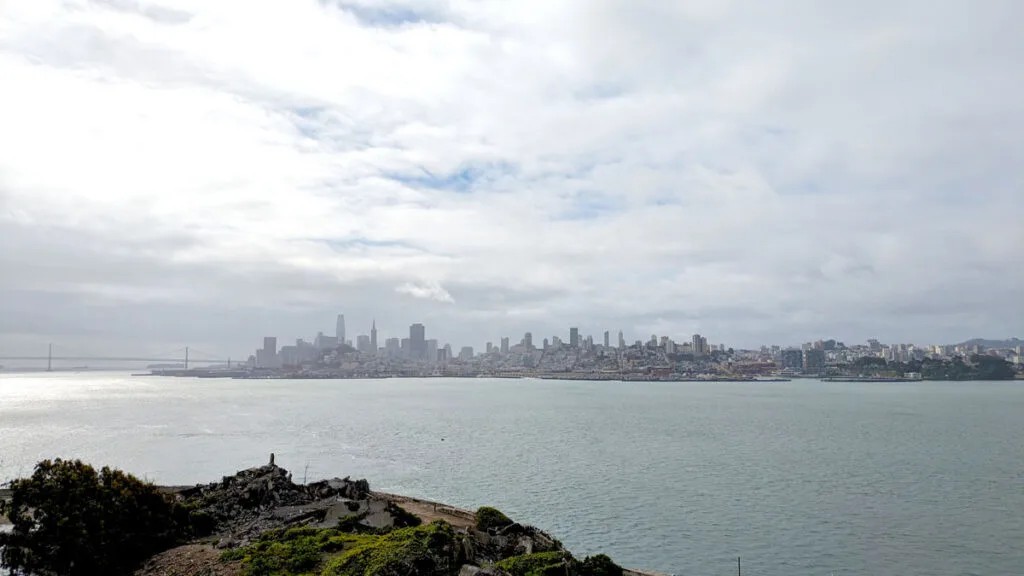 a slightly foggy skyline view of san francisco but there's some sun peaking through adding some yellow and brightiness. It's across the bay from alcatraz island.