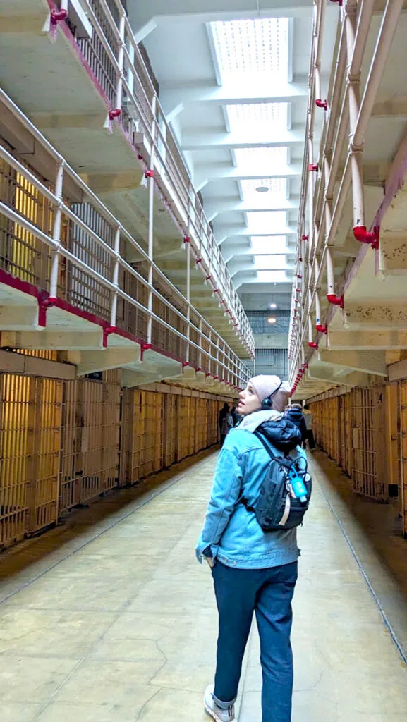 a woman walks forward in the cellblock of alcatraz. There are jail cells on either side of the hallway, and there are three levels . the ceiling has sun roofs letting in some much needed light to the cell block.