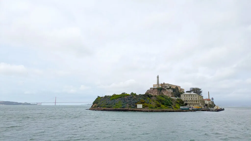 a sole island is in the middle of choppy water: alcatraz. There's a lighthouse and two buildings in the middle of the island. In the distance you can see the red golden gate bridge among the fog. The sky is overcast and grey.