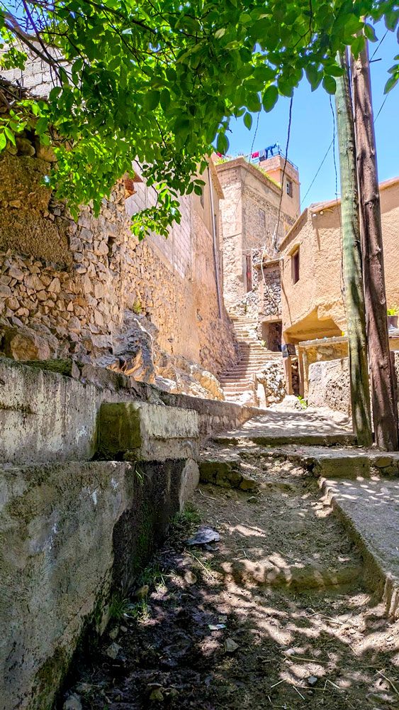 walking through a Berber Village in the High atlas Mountains Morocco. There's a paved road with a dir road next to it leading up into the village. A stone rock wall separates you from the rest of the village. There are stairs leading up to a large building, on the rooftop are some clothes drying on hangers. There are even wires that are going through the village. 