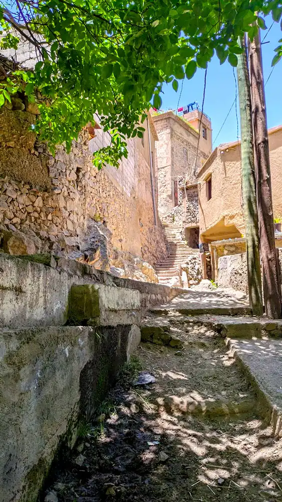 walking through a Berber Village in the High atlas Mountains Morocco. There's a paved road with a dir road next to it leading up into the village. A stone rock wall separates you from the rest of the village. There are stairs leading up to a large building, on the rooftop are some clothes drying on hangers. There are even wires that are going through the village.