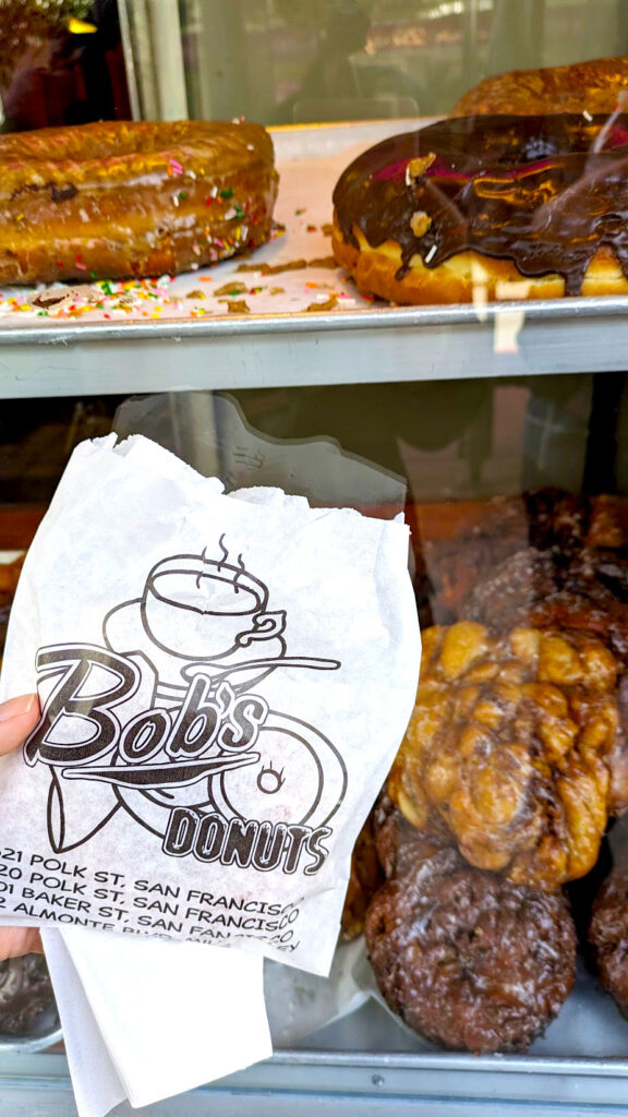 A bag of Bob's Donuts with the location addresses. Behind the bag are their donuts on display, including two GIANt donuts with chocolate glaze, below are their apple fritters.