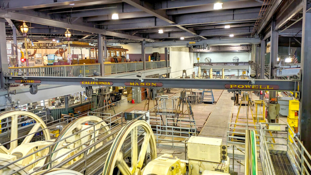 inside the cable car museum seeing all the cables from the cable cars being pulled by giant wheels and going across what looks like a warehouse. There's a beam above the wheels with signs for Hyde, California, Mason, and Powell, it shows which cables are going to which route.
