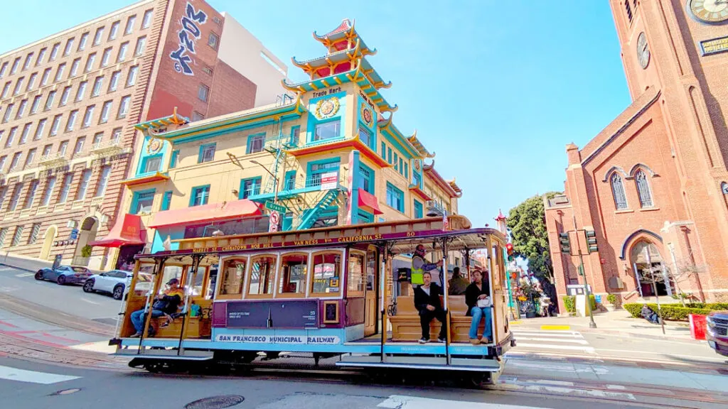 a cable car drives by Chinatown in San francisco. You know it's chinatown because the building in the background is a traditional chinese architecture roof. It's painted a soft yellow, teal, and red highlights. The cable car only has a few people sitting on the benches facing outwards.